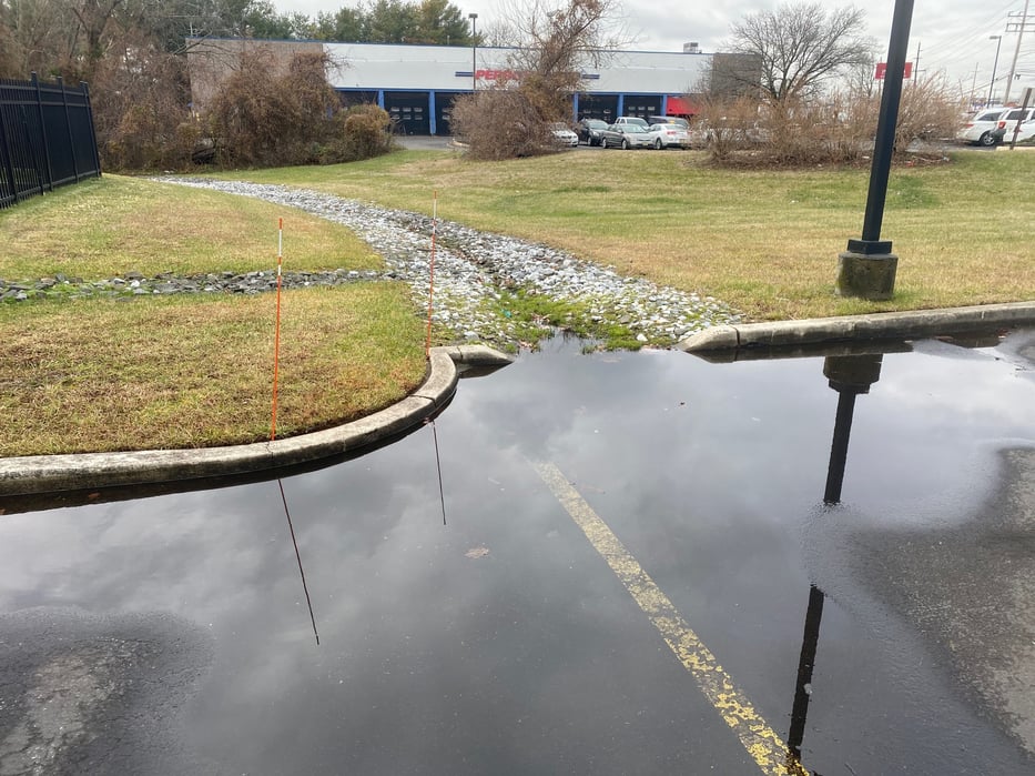 Clogged rip rap drainage swale with grass and debris preventing water flow, causing pooling in Toms River commercial lot.